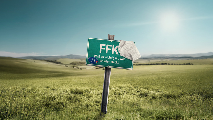 Ein grünes Schild mit der Aufschrift "FFK", an dem Unterwäsche hängt, steht in einer sonnigen, grasbewachsenen Landschaft. - © PMT 2025