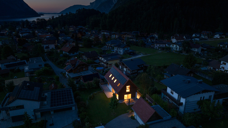 Ein modernes Haus mit warmer Beleuchtung, umgeben von dunklen Häusern und Bergen in der Abenddämmerung. - © Fronius