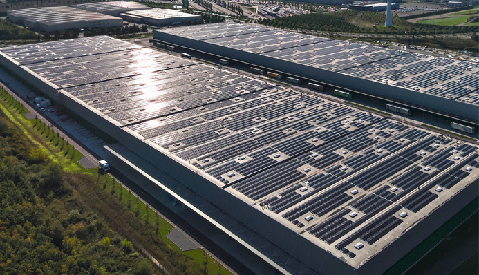 Luftaufnahme eines großen, mit Solarmodulen bedeckten Lagerhausdachs, umgeben von Grün. - © Foto: Resource