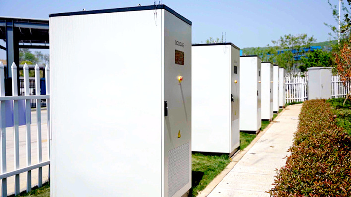 Mehrere weiße Schaltschränke für den Außenbereich reihen sich an einem sonnigen Tag neben einem Gehweg und einem weißen Zaun auf. - © Goodwe