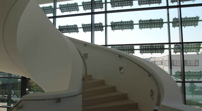 Eine Wendeltreppe mit geschlossener Brüstung vermittelt den Eindruck von Galerie oder Theater. Überall in dem Atrium ist die Photovoltaikanlage vor der Gasfassade allgegenwärtig.